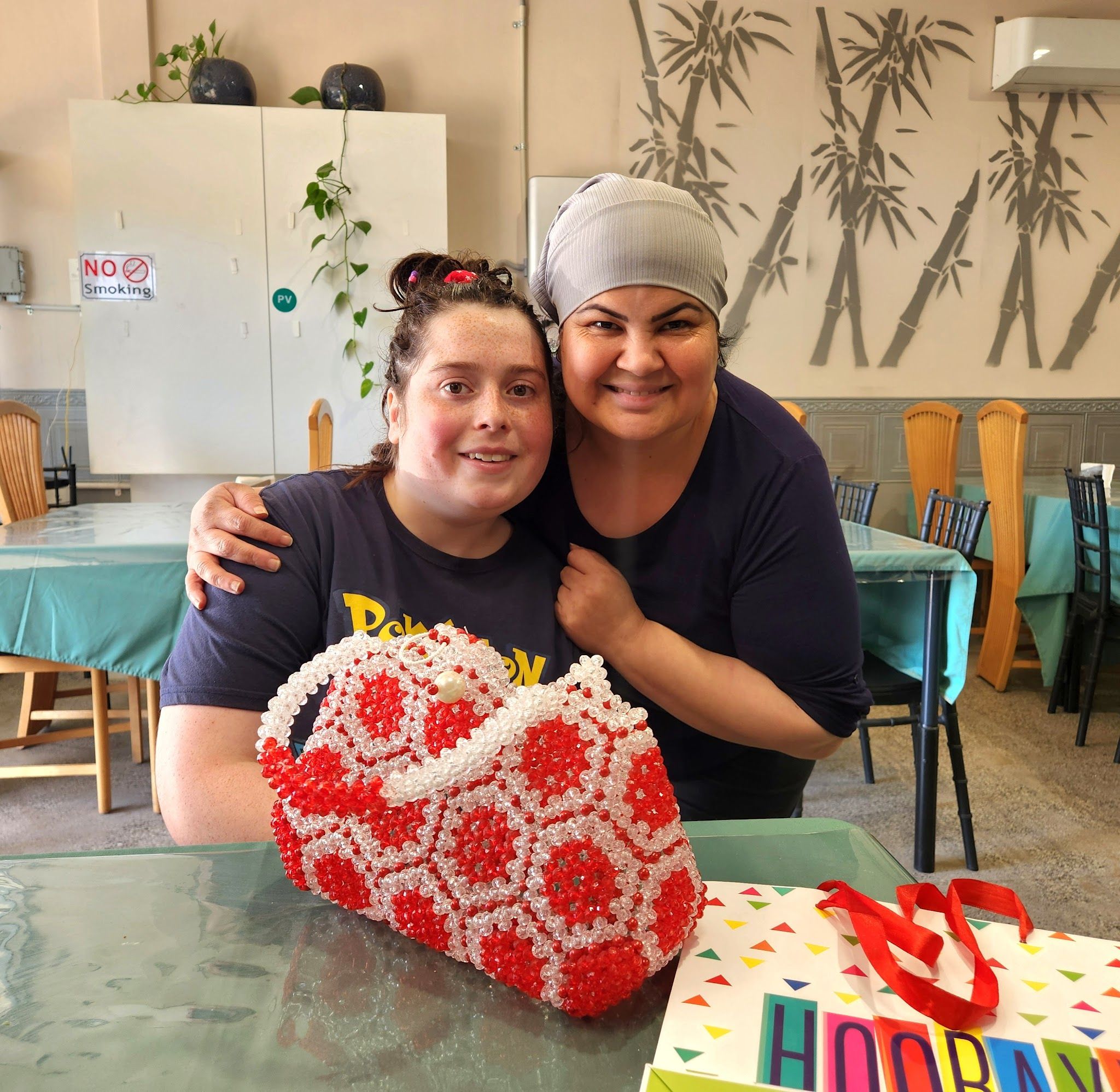Two women smiling at a table in the dining room with bamboo mural wallpaper and teal-covered tables at Nad's